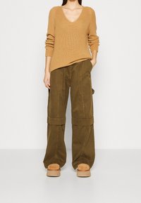 Maglione a coste color sabbia con scollo a V, abbinato a pantaloni cargo verde oliva a gamba larga con ampie tasche e orli piegati. Scarpe platform beige morbide.