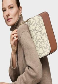 Suéter beige con cuello acanalado, modelo sosteniendo un bolso bicolor; un lado en crema con diseño texturizado, el otro lado de cuero marrón con correa de cadena.