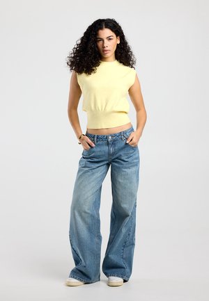 Frau mit lockigem Haar, die ein ärmelloses gelbes Oberteil und weit geschnittene blaue Jeans trägt, steht mit den Händen in den Taschen vor einem einfarbigen Hintergrund.