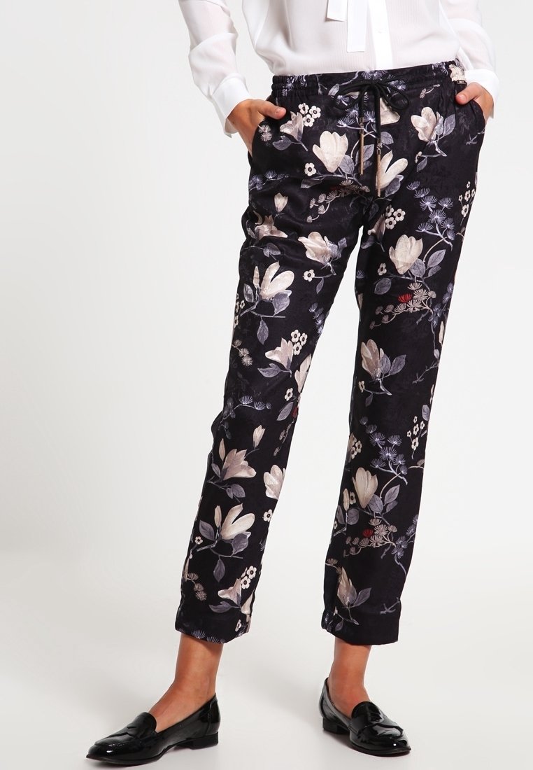 Personne portant un pantalon noir à imprimé fleuri, un chemisier blanc et des mocassins noirs, debout les mains dans les poches devant un fond uni.