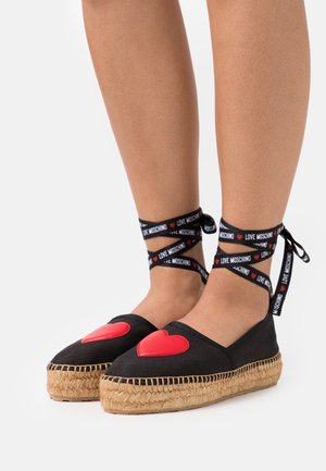 Espadrilles noires avec appliqué en forme de cœur rouge, semelle en jute et sangles noires portant le texte "LOVE MOSCHINO". Design plat et style décontracté.