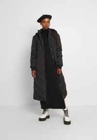 Lång svart puffer-jacka med huva och dragkedja, med diagonala quiltade mönster. Matchad med en svart klänning och vita snörstövlar.