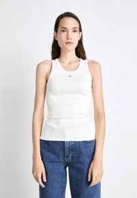 White ribbed tank top with a round neckline and a small black "ck" logo at the center. Paired with blue high-waisted jeans.