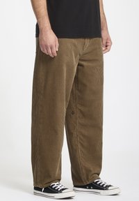 Brune corduroy bukser med brede ben, der har subtile lodrette striber, en tekstureret overflade og et logodetalje på venstre side. Sorte sneakers.