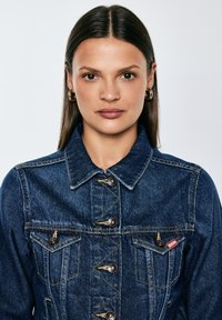 Jauna moteris su tiesiais tamsiais plaukais, dėvinti auksinius lankinius auskarus ir tamsiai mėlyną džinsinę striukę su sagomis, žiūrinti tiesiai į priekį priešais paprastą foną.