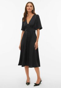 Vestido negro con escote en V, mangas cortas con volantes y cierre frontal con botones. Tejido liso y fluido que cae hasta la mitad de la pantorrilla. Se lleva con tacones negros.