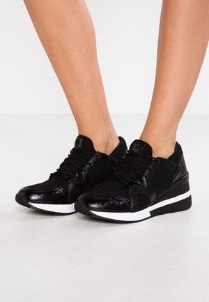 Czarne sportowe sneakersy z białymi podeszwami noszone na nogach, z siateczkową tkaniną i błyszczącymi teksturowanymi akcentami, z zawiązanymi sznurowadłami.