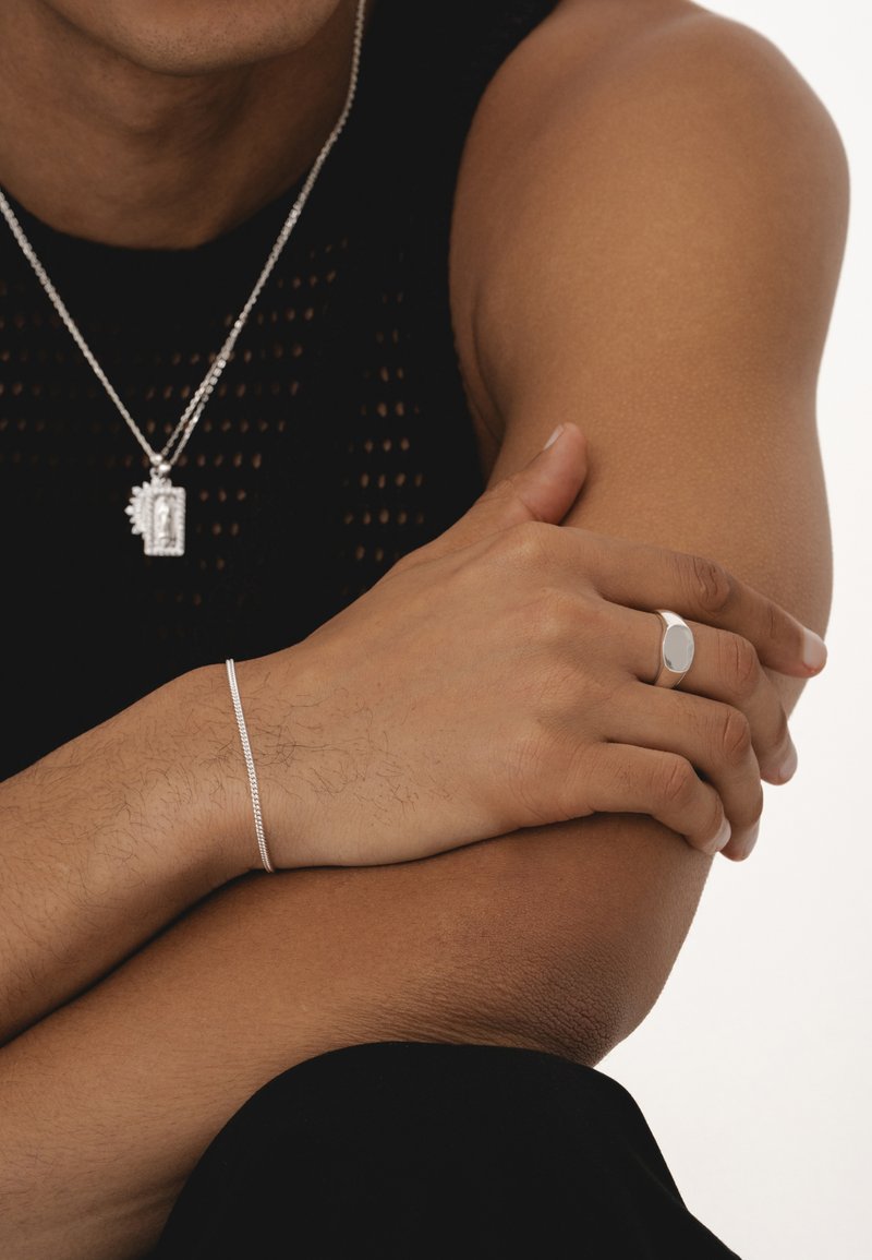 Bague en argent avec une face ovale, bracelet en argent fin et collier en chaîne argentée à plusieurs couches avec un pendentif rectangulaire. Haut sans manches texturé noir.