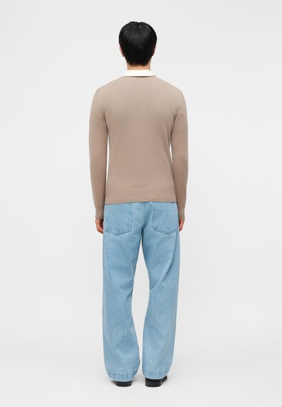 Άτομο που στέκεται με την πλάτη προς τα εμπρός, φοράει μπεζ πουλόβερ με μακριά μανίκια, ανοιχτό μπλε φαρδιά τζιν και μαύρα παπούτσια σε απλό φόντο.