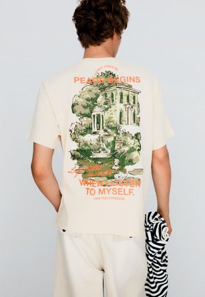 Osoba nosząca kremowy t-shirt z zieloną ilustracją ogrodu i budynku oraz pomarańczowym napisem: „Pokój zaczyna się, gdy słucham siebie."