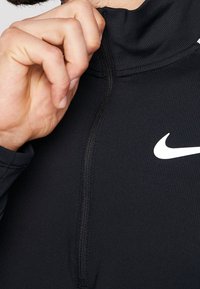 Czarny sportowy polar z wysokim kołnierzem, żebrowaną teksturą, zamkiem z przodu oraz białym logo Nike na piersi. Zbliżenie na obszar szyi.