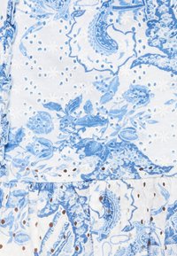 Bílá látka s modrými květinovými a paisley vzory, zdobená detaily v podobě dírek a texturovaným povrchem. Volánové okraje zvýrazňují design.
