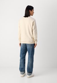 Kreminė sportinė palaidinė su tekstūruotu paviršiumi, su raukšlėtais rankogaliais ir apačia, derinama su šviesiai mėlynomis šiek tiek nusidėvėjusiomis plačiomis kelnėmis ir baltais sportbačiais.