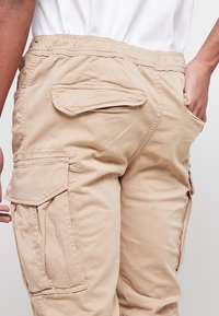 Le pantalon cargo beige présente de multiples poches, une taille élastique et une coupe décontractée. Le tissu semble léger avec une texture lisse.