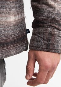 Bruine en grijze gebreide langemouwen shirt met een zichtbare naad en een klein label op de manchet. De stof vertoont een subtiel gestreept patroon.