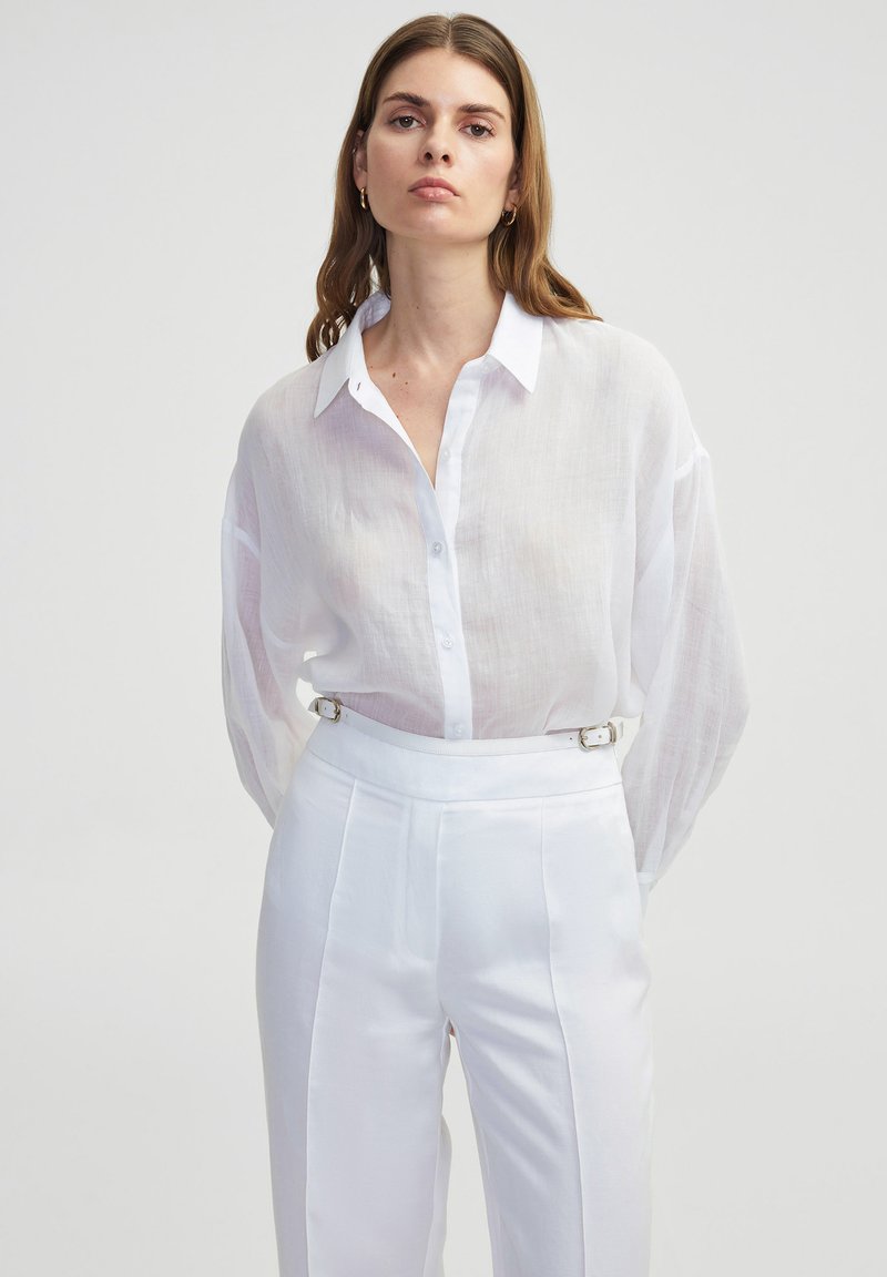 Camicia bianca, leggera e semi-trasparente con maniche lunghe e colletto, abbinata a pantaloni bianchi a vita alta con dettagli in cintura.