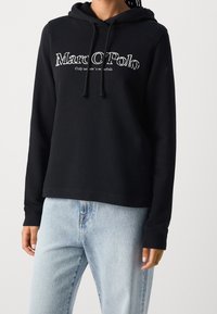 Sudadera con capucha negra de tela suave con un corte relajado, con un logo frontal en blanco y cordones en la capucha. Combinada con vaqueros azul claro.