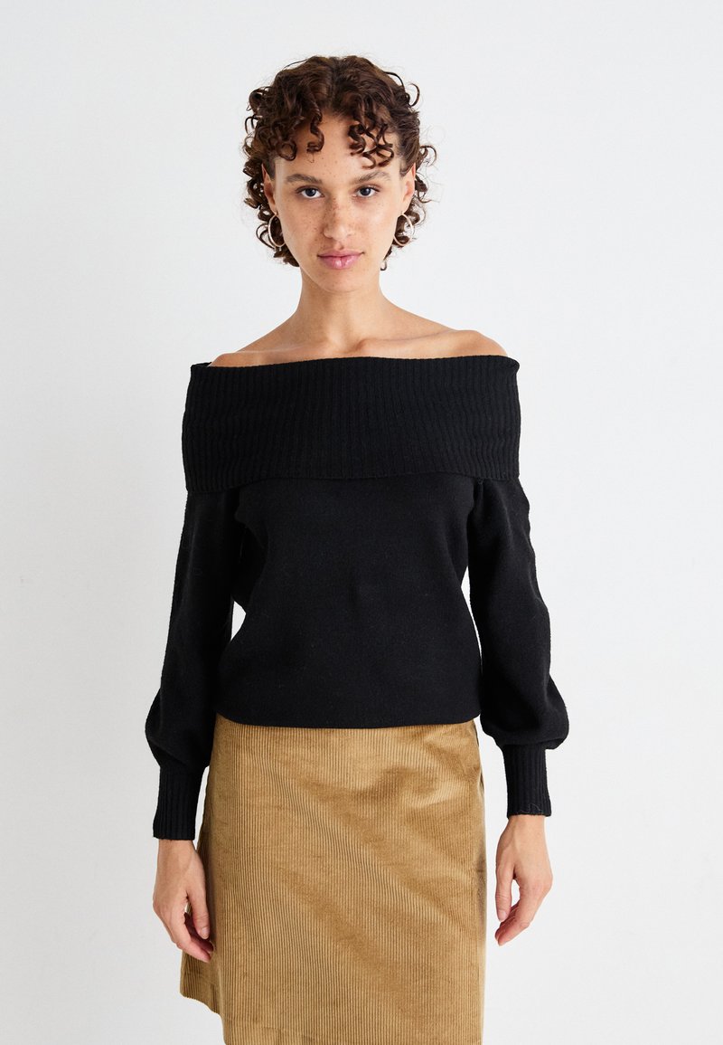 Off-the-shoulder svart strikkegenser med ribbet halslinning, lange puffede ermer og tettsittende kropp, kombinert med en beige cordfløyelskjørt.