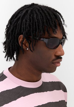Uomo con dreadlocks corti che indossa occhiali da sole neri, un piccolo orecchino a bottone e una camicia a righe rosa e grigio scuro che guarda di lato.