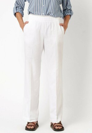 Persona che indossa pantaloni bianchi con vita elastica con le mani nelle tasche, abbinati a una camicia a righe blu e bianche e sandali marroni.