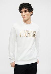 Νέος με σγουρά μαλλιά που φοράει λευκό φούτερ με χρυσά γράμματα "Versace Jeans Couture", κοιτάζοντας προς τα πλάγια.