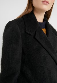 Abrigo de lana negro texturizado con cuello de muesca y un acabado suave, que presenta un diseño ajustado que cubre el hombro y la parte superior del brazo.