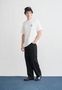 Bílé oversized tričko s malým fialovým logem, černé široké kalhoty a černý klobouk typu bucket, doplněné černou obuví s bílými detaily.