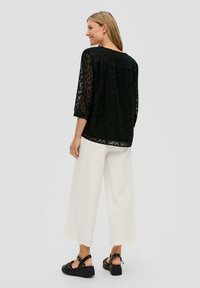 Zwarte doorschijnende blouse met een textuur in luipaardpatroon, 3/4 mouwen, gecombineerd met brede witte broek en zwarte chunky sandalen.