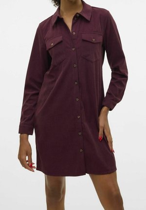 Kobieta ubrana w długą, zapinaną na guziki sukienkę z ciemnofioletowego sztruksu z kieszeniami na piersi, stojąca na tle jednolitego białego tła.