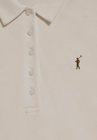 Vit polo-shirt i texturerad tyg med tre knappar fram. Har en liten broderad brun figur som håller en golfklubba.