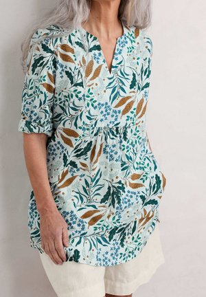 Kvinde med gråt hår iført en blomstret bluse med lange ærmer, der er rullet op, og lyse shorts, stående mod en ensfarvet væg.