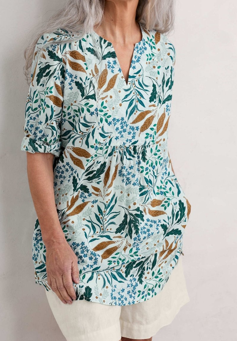 Femme aux cheveux gris portant un chemisier à manches longues à motif floral avec manches retroussées et un short de couleur claire, debout contre un mur uni.
