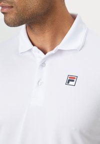 Biely polo tričko s textúrovanou látkou, dve gombíky pri golieri, s malým viacfarebným logom na ľavej hrudi.
