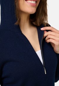 Haut à capuche zippé bleu marine avec une texture douce, doté d'une capuche, d'une fermeture éclair argentée et d'un bord côtelé. Gros plan autour du col et du visage.