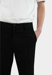 Schwarze Hose mit glatter Textur, Knopfverschluss, seitlichen Taschen und taillierter Passform, kombiniert mit einem weißen Kurzarmhemd.