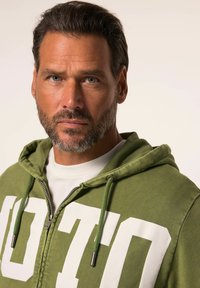 Sudadera con cremallera de color verde oliva con las letras blancas "OTO" en la parte delantera, que cuenta con capucha ajustable y tejido texturizado.