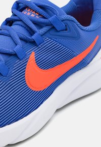 Chaussure de sport Nike avec une tige texturée bleue, logo swoosh orange, semelle blanche plate et lacets bleus. Les détails incluent des rayures horizontales.