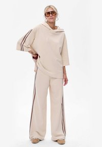Beżowa bluza z kapturem o szerokich rękawach, zestawiona z beżowymi spodniami z szerokimi nogawkami z ciemnoczerwonymi paskami po bokach. Modelka w okularach przeciwsłonecznych.
