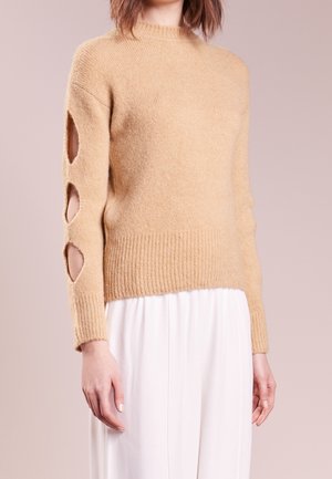 Un pull beige, doux, avec un ourlet côtelé et un col arrondi. Présente des détails découpés au niveau des manches. Associé à un pantalon blanc.