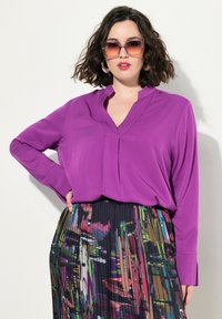 Blusa viola a maniche lunghe con scollo a V e pieghe, abbinata a una gonna plissettata con un motivo astratto multicolore.