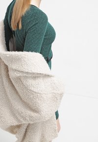 Vihreä neulepaita, jossa on tekstuuri, kerroksittain paksun kermaisen takin alla, jossa on pehmeä, pluuma päällinen ja avoin etuosa.