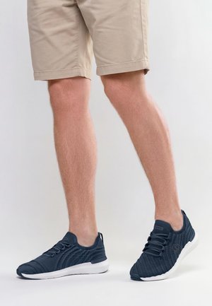 Jambes portant un short beige et des baskets en tricot bleu marine à semelles blanches, debout sur un fond clair uni.