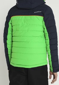 Personne portant une veste matelassée verte et noire avec capuche, logo "Dare 2b" dans le haut du dos, debout dos à la caméra.