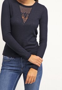 Haut marine à manches longues côtelé avec un détail en triangle en dentelle à l'encolure, associé à un jean en denim. Le tissu semble texturé et ajusté.