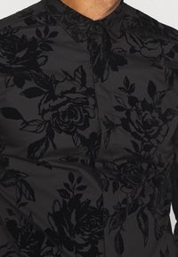 Zwarte bloempatronen blouse met een soepele stoftextuur, een kraag en knoopsluiting. Het ontwerp heeft verhoogde, fluwelige rozen en bladeren.