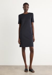 Robe marine à manches courtes avec un col rond, silhouette ajustée, poches latérales et une subtile accent métallique. Portée avec des mules noires à bout ouvert.