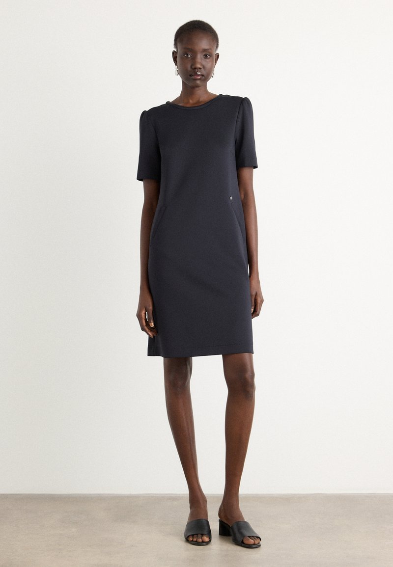Robe marine à manches courtes avec un col rond, silhouette ajustée, poches latérales et une subtile accent métallique. Portée avec des mules noires à bout ouvert.