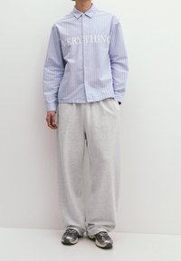 Ljusblårandig skjorta med knappar som har vit text "EVERYTHING," i kombination med grå sweatpants och mörka sneakers. Enkelt mönster, avslappnad passform.