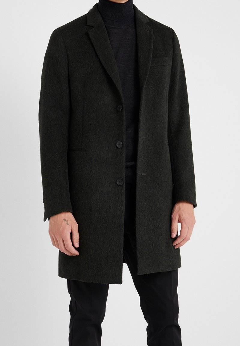 Abrigo de lana de color verde oscuro con cuello muesca, botones en la parte delantera y un bolsillo en el pecho izquierdo. Corte entallado y textura suave.