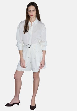 Kobieta nosząca białą, zapinaną na guziki koszulę oraz białe szorty z wysokim stanem i paskiem, ozdobione drobnym haftem, zestawione z czarnymi, niskimi mules.